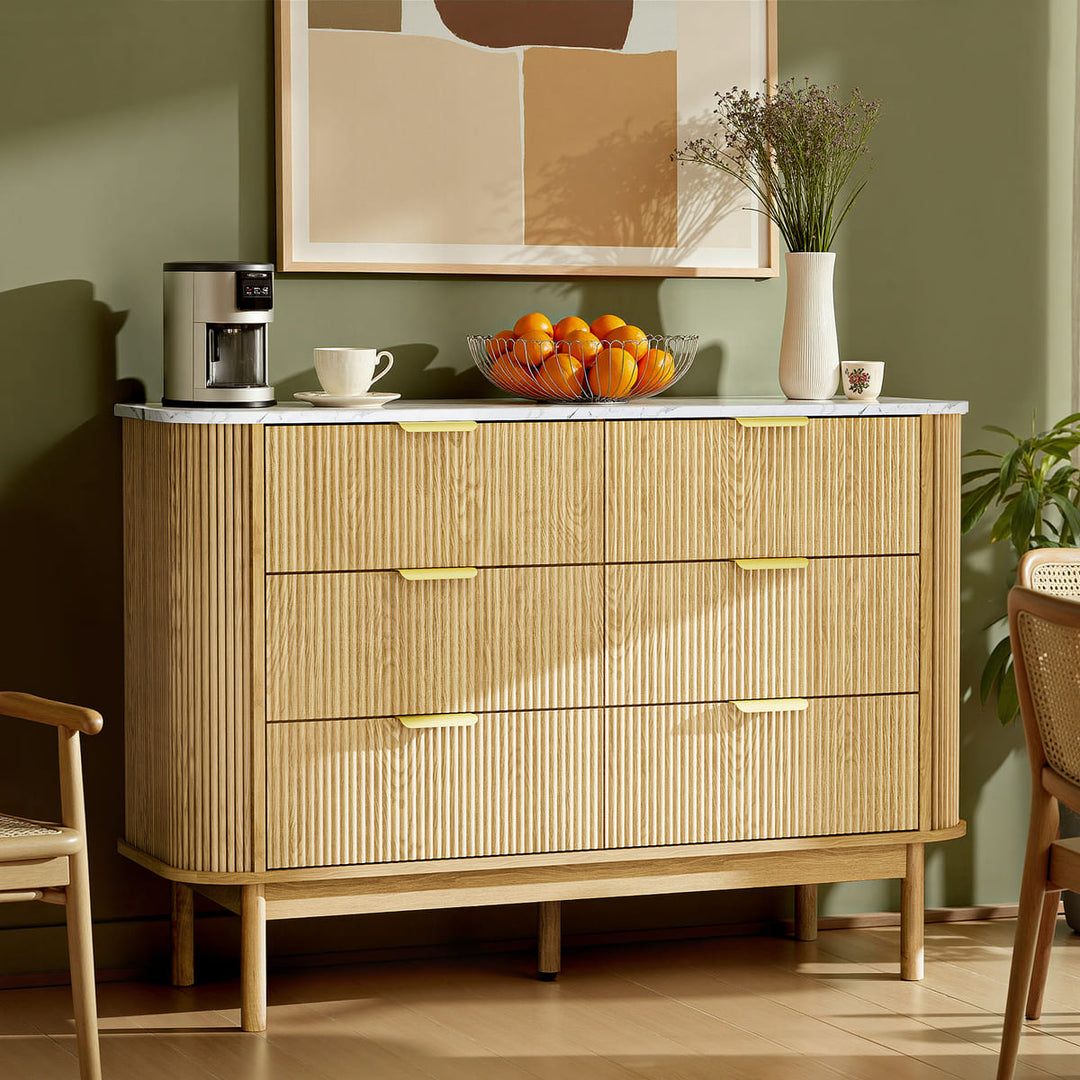 Wooden dresser with drawers, coffee maker, and fruit bowl in a room with a painting on the wall.