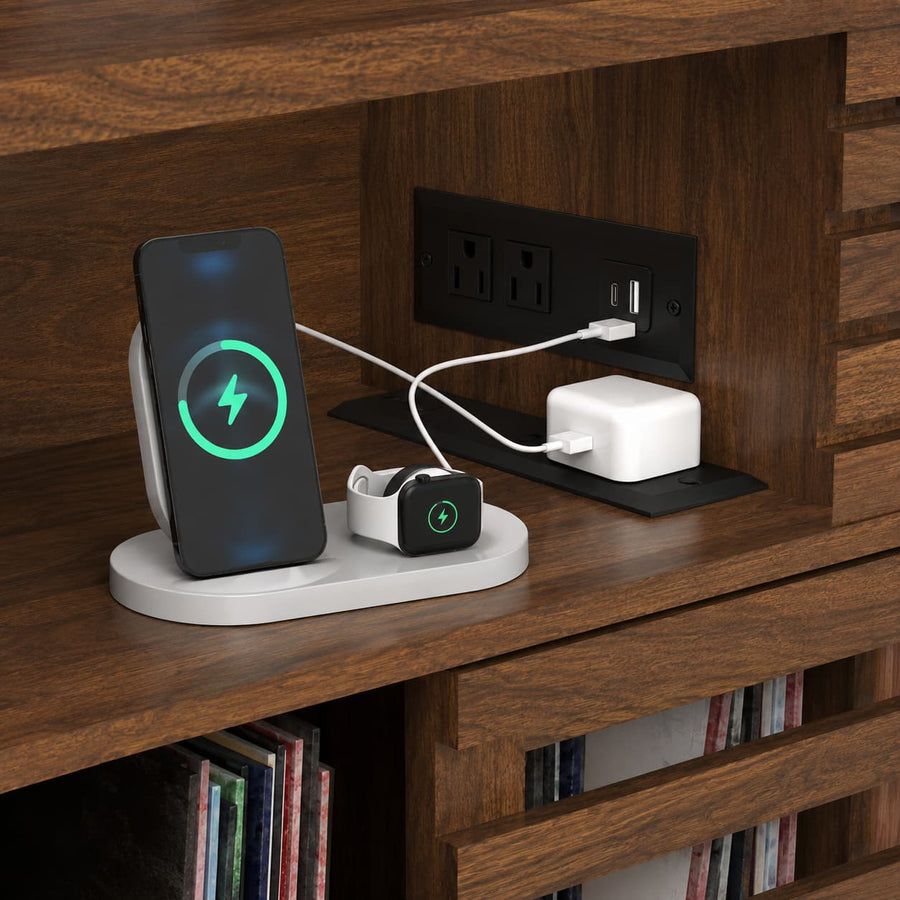 A white multi-device wireless charging stand sits on a dark wood shelf, charging a smartphone, a smartwatch, and a white power adapter. The stand is shown in a home environment next to wall outlets.